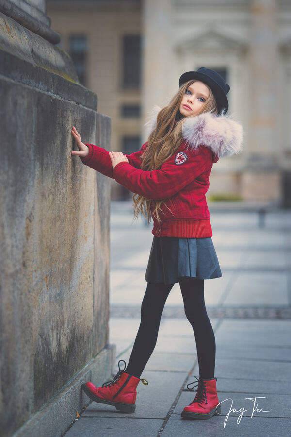 Young girl with long blonde hair, red jacket, red shoes and a black hat.