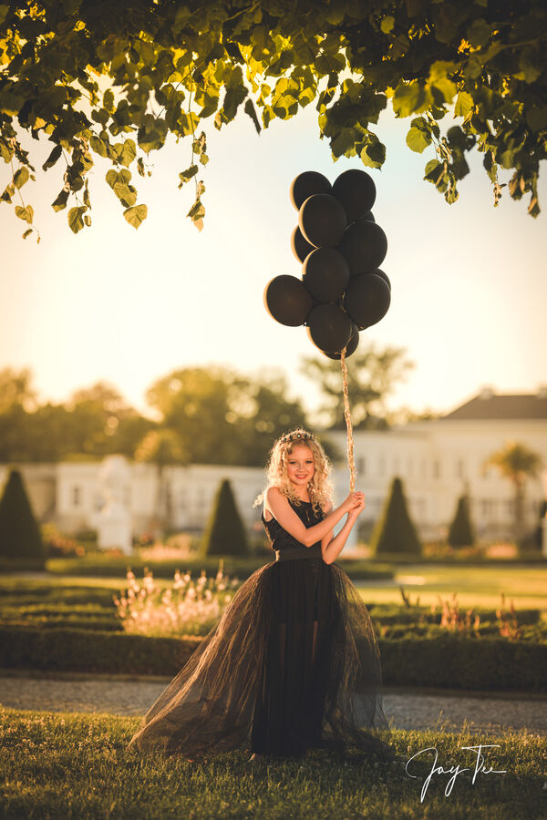 Little Girl with a black dress and black balloons at sunset in a royal garden.